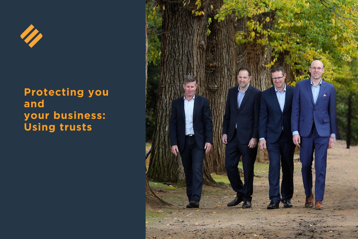 Protecting you and your business using trusts Four professionally dressed male advisers from Johnsons MME walk side by side through a tree-lined park, representing trust and strategic business advice.